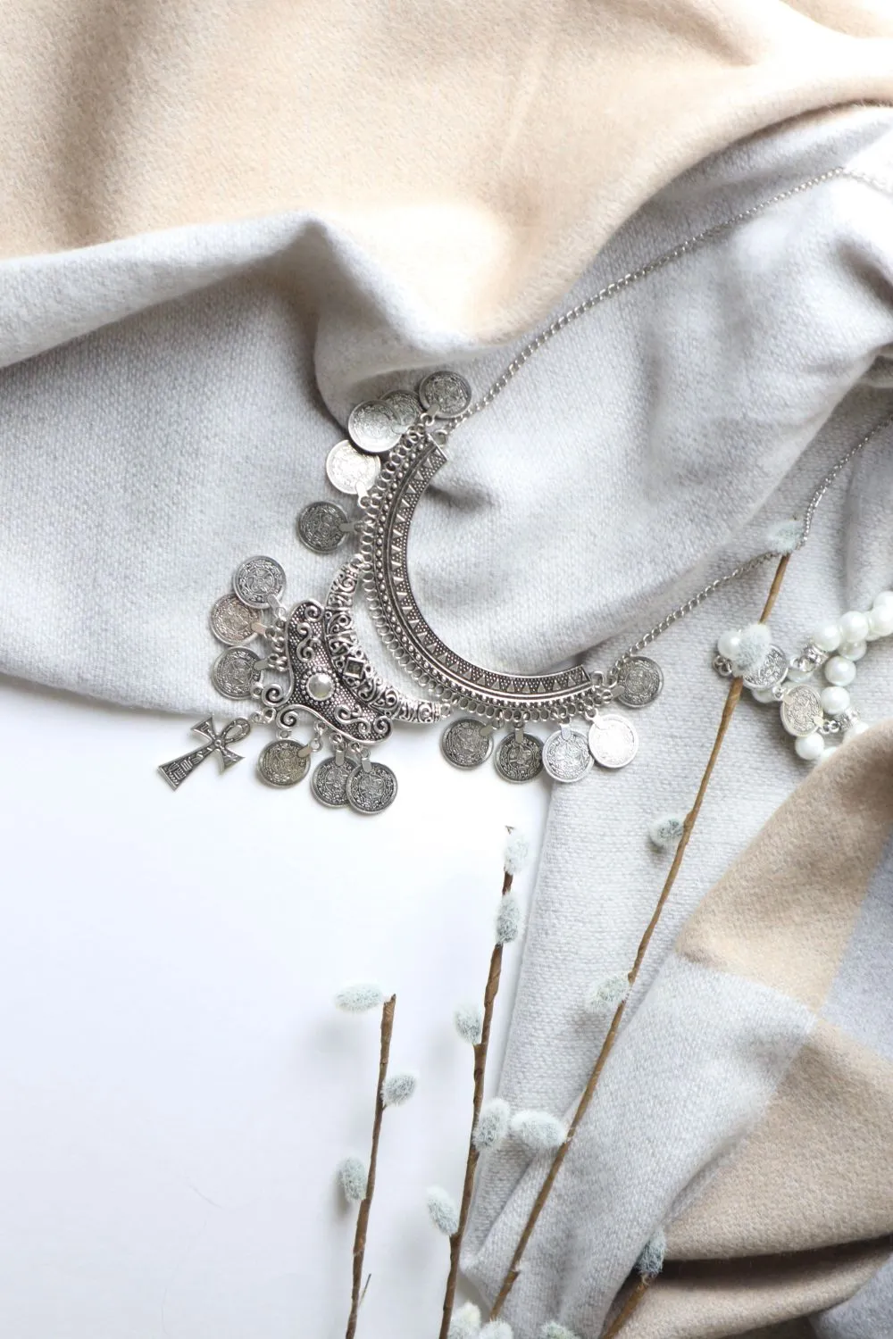 A silver necklace of stamped discs and coins laid across a soft oatmeal knitted throw.