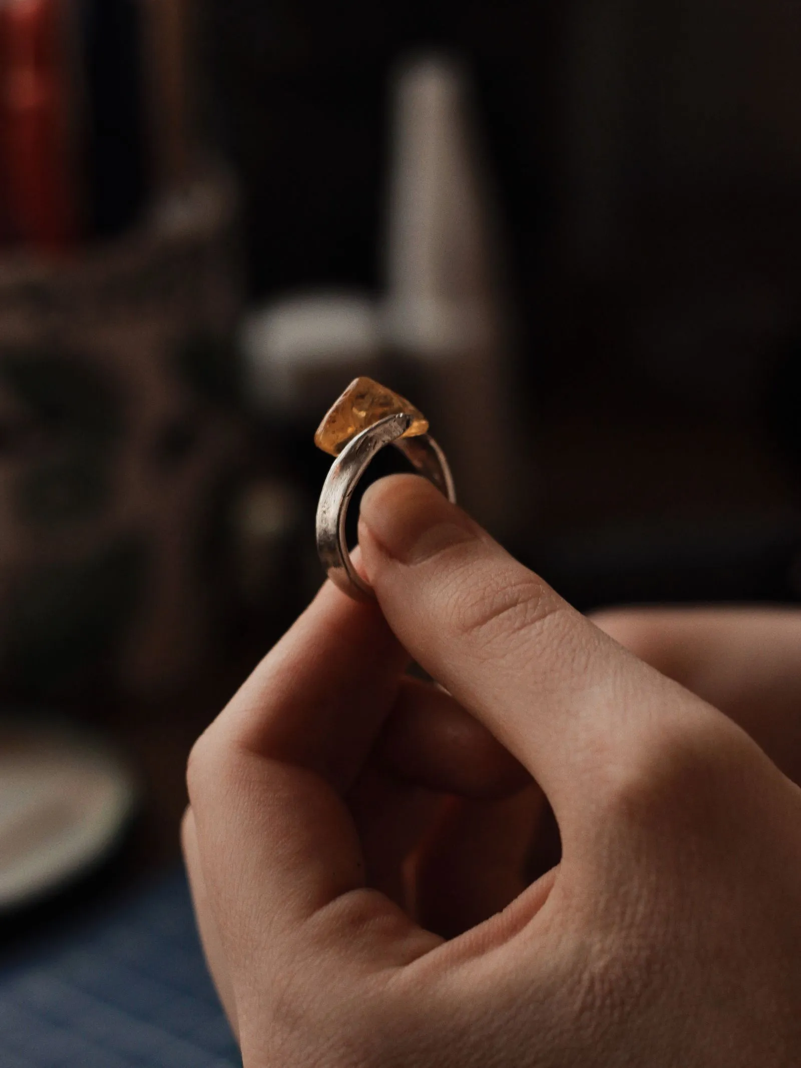 A hand holding a finished silver ring with an amber-coloured stone, in warm bench light.