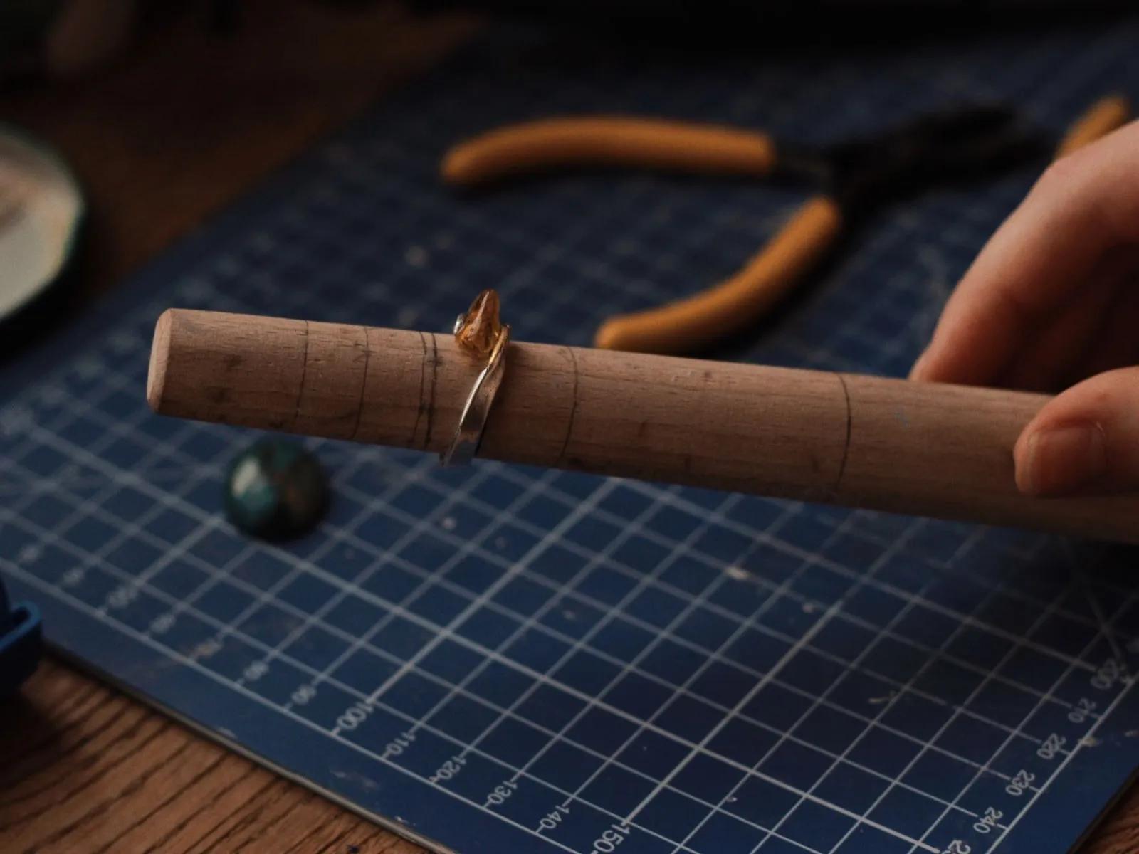 A silver ring resting on a wooden mandrel beside pliers, on the blue cutting mat of a jeweller's bench.