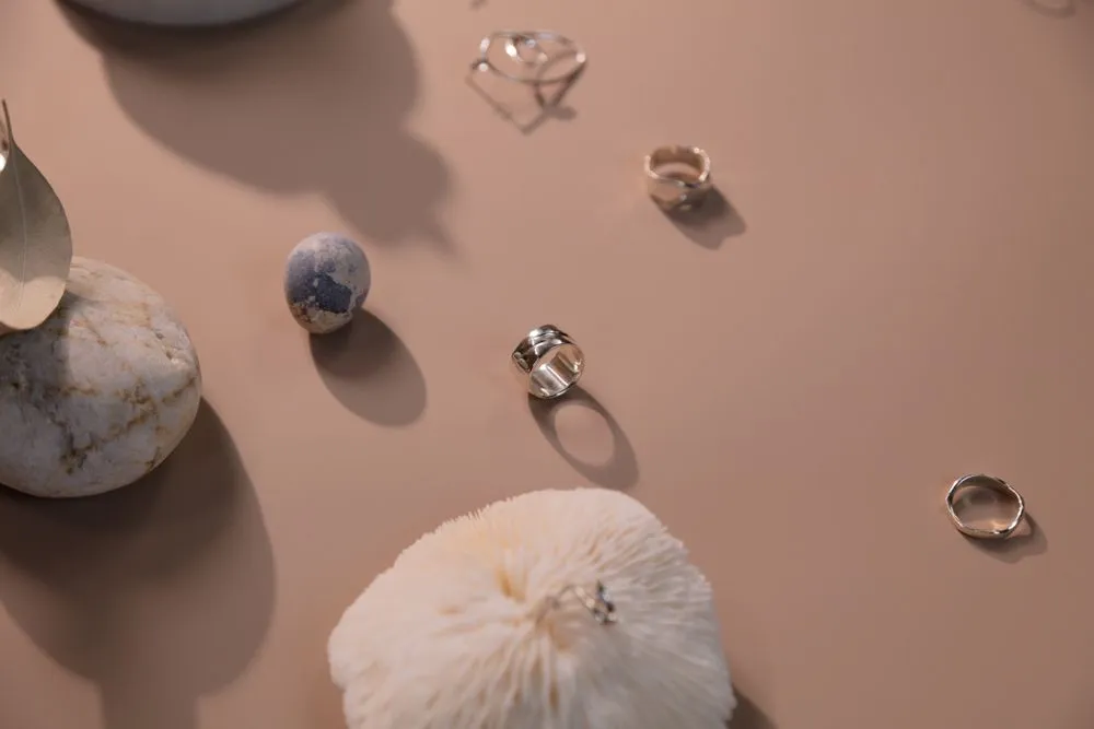 Three slim silver rings scattered on a sand-coloured ground with pebbles and a shell.