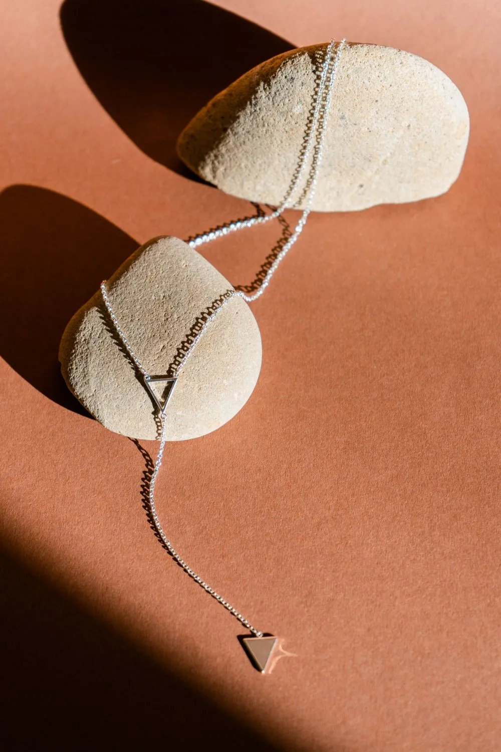 A small silver triangle pendant on a fine chain, laid over pale river stones on a terracotta ground.
